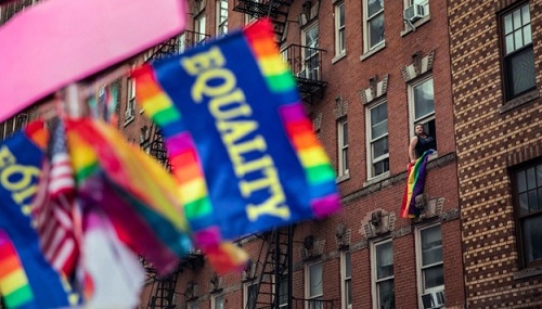 Equality rainbow flag in foreground with apartment building behind and person hanging out window waving their own flag