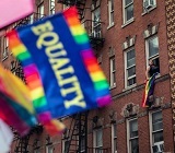 Equality rainbow flag in foreground with apartment building behind and person hanging out window waving their own flag