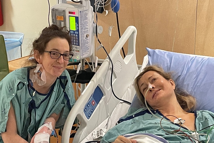 Bobbi and Robin in hospital gowns, holding hands, as Bobbi lies in hospital bed and Robin sits at her side