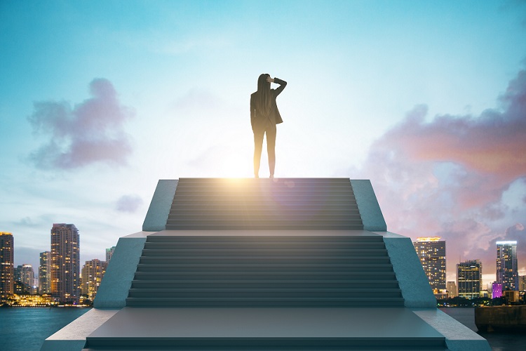 Young women in suit atop wide stairs looking out over at sunny city skyline