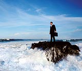 man in a suit standing on a rock in middle of lake, waves crashing around him