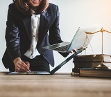 Photo of young female attorney preparing notes at table in court with law books, laptop and scales of justice