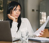 Woman seated at desk with laptop, viewing printed document with confusion