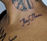 Side image of a person's upper torso and neck, featuring many black ink tattoos, the most prominent of which is the words "They/Them" in cursive writing