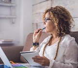 A woman sits on a couch with her laptop on her lap, a notepad in her, and his the tip of the pencil in her mouth as she gaze into the distance in deep thought