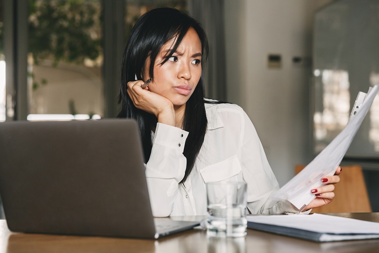 Professional woman seated at desk with laptop, reading printed document with confused expression