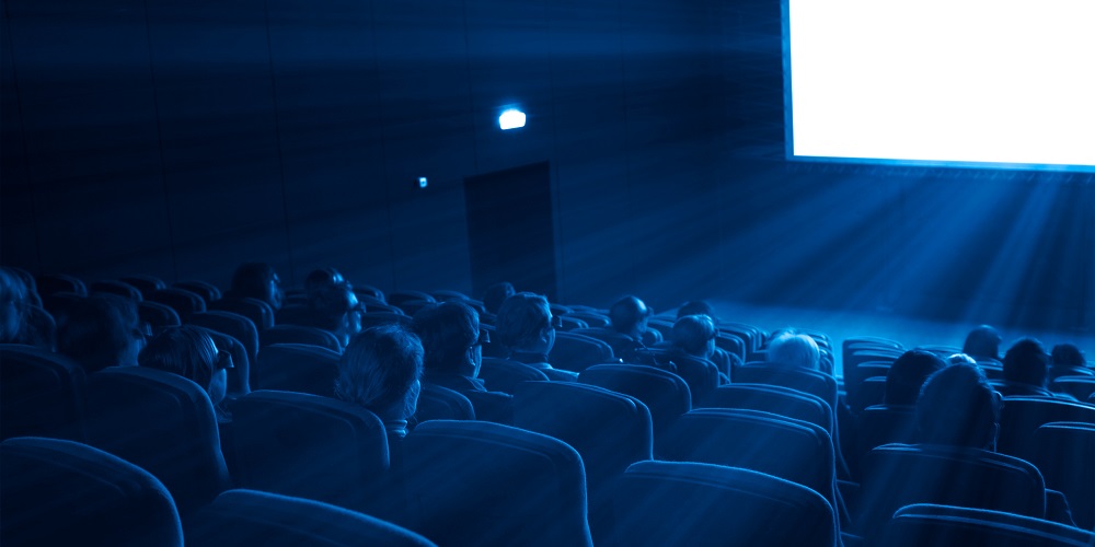 blue-toned photo of movie theatre with audience facing brightly lit screen