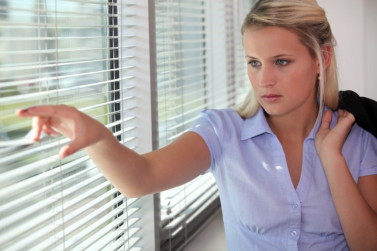 woman peering out nervously from behind horizontal blinds in her office