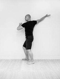 man in exercise gear leaning his right side against the wall with right arm stretched out behind him, bracing himself against wall with left hand