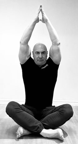 man in exercise gear sitting cross-legged on floor with arms stretched up, palms together