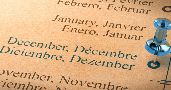Close-up of a multilingual calendar showing the months July and August in English, French, Spanish, and German, with a blue pushpin marking Decembber