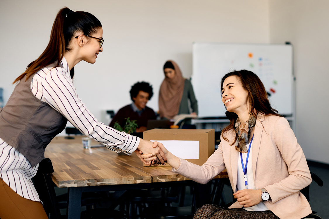 Two professional women shaking hands in an office setting, with colleagues working in the background.
