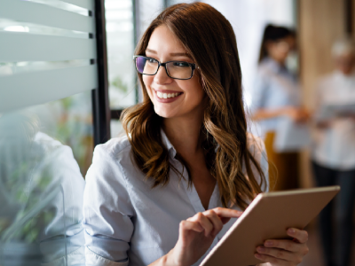 Smiling woman with glasses using a digital tablet in a bright office setting, with colleagues conversing in the background.