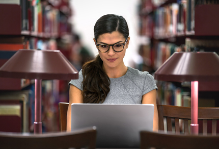 Woman with long dark hair and glasses sitting at a library desk, working on a laptop, surrounded by bookshelves and desk lamps, in a quiet study environment.