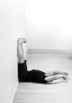 man lying on floor in exercise gears with legs bent upward at a 90 degree angle, resting on wall, and arms stretched back behind head