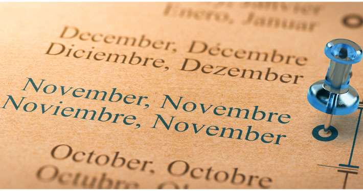Close-up of a multilingual calendar showing the months July and August in English, French, Spanish, and German, with a blue pushpin marking November