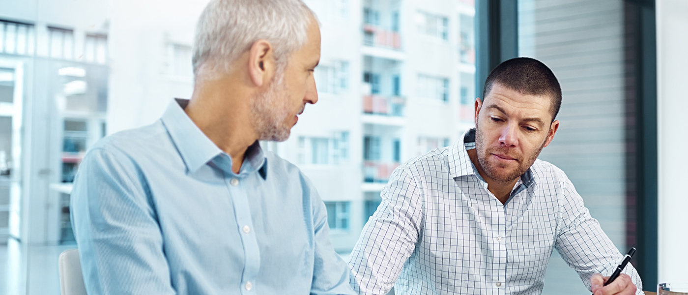 The image appears to show two men engaged in a discussion in a professional setting, possibly an office, with large windows and a cityscape in the background. Let me know if you need a detailed descri