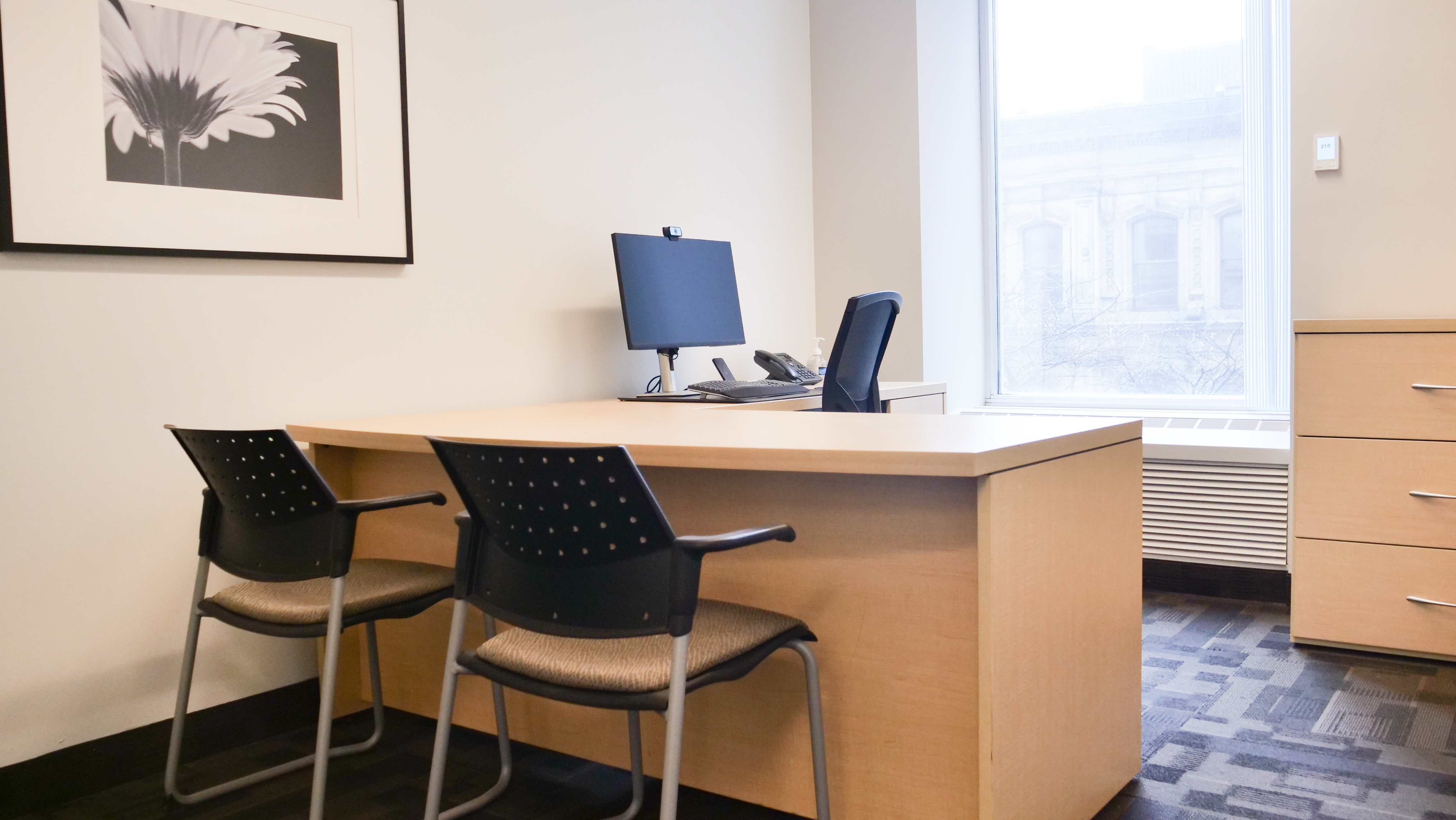 Minimalist office corner with wooden desk, black chairs, and mounted cabinet in a bright modern workspace.