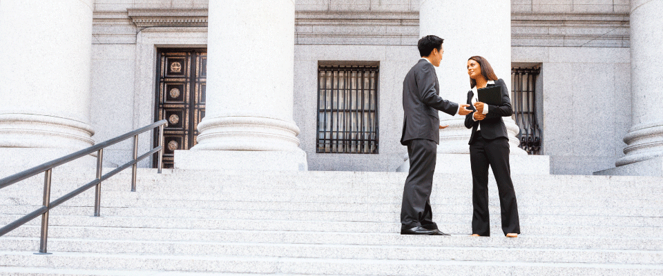 Two business professionals engaged in conversation on the steps of a grand courthouse, symbolizing legal discussion or professional collaboration.