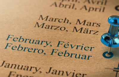 Close-up of a multilingual calendar showing the months July and August in English, French, Spanish, and German, with a blue pushpin marking February