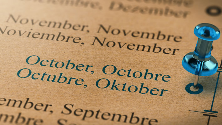 Close-up of a multilingual calendar showing the months July and August in English, French, Spanish, and German, with a blue pushpin marking October.