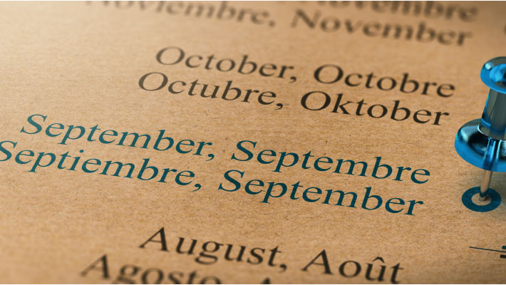 Close-up of a multilingual calendar showing the months July and August in English, French, Spanish, and German, with a blue pushpin marking September