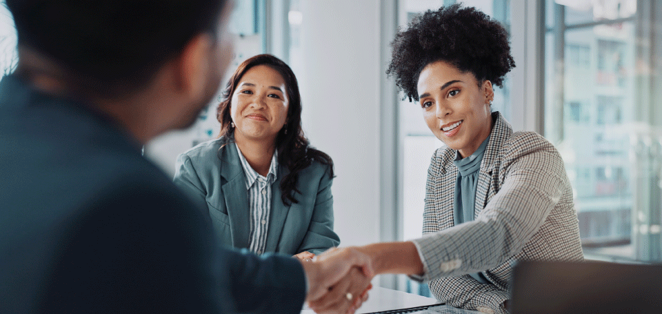 Two professional women in a business meeting, smiling as one extends a handshake to a colleague, symbolizing agreement and collaboration.