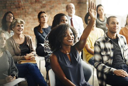 Diverse group of adults seated in a classroom or seminar setting, with a woman in the front center raising her hand to participate.