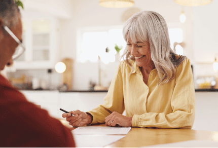 Smiling older woman with gray hair signing documents at a kitchen table while talking with another person.